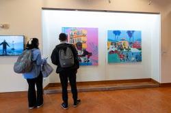 Two visitors stand and observe a colorful painting behind glass.